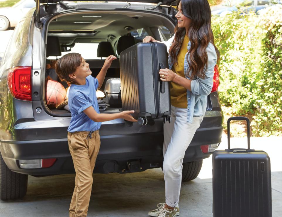 A mom and her child lifting a heavy suitcase out of the trunk of an SUV
